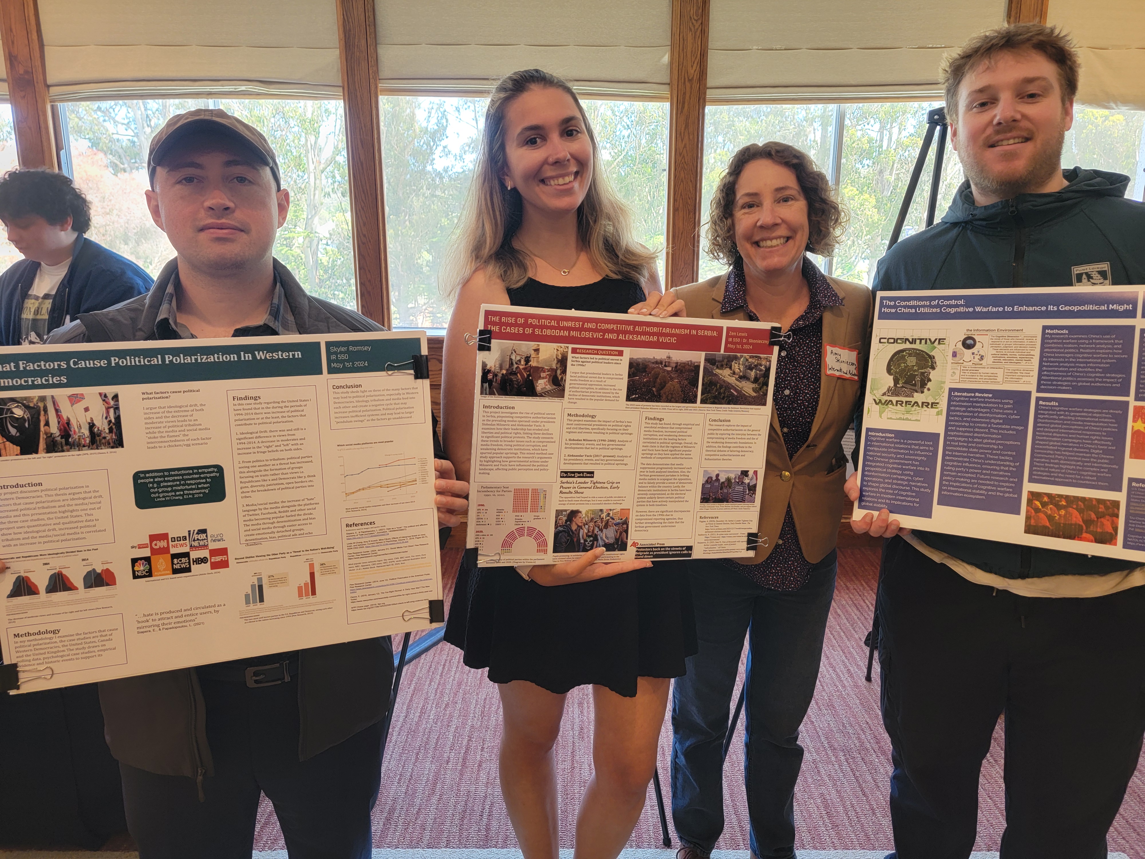 Three students from IR 550 stand and face the camera. They are smiling and holding their presentation posters. Professor Skonieczny stands just behind them, also smiling to the camera.