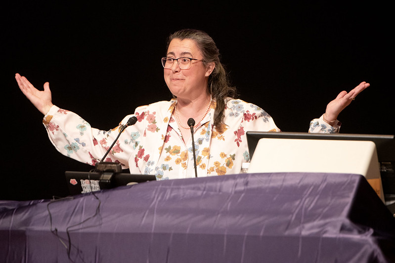 Professor Ellis gestures, holding both arms out, while speaking from behind a podium. She wears a white floral blouse.