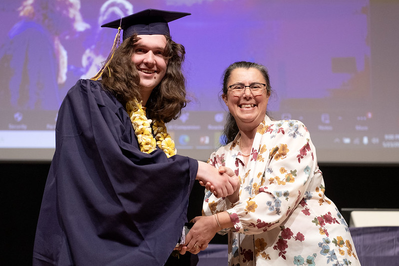 Lexy Johnstone and Burcu Ellis face the camera while shaking hands. Lexy wears purple graduation regalia and a yellow lei. Professor Ellis wears a white floral blouse.