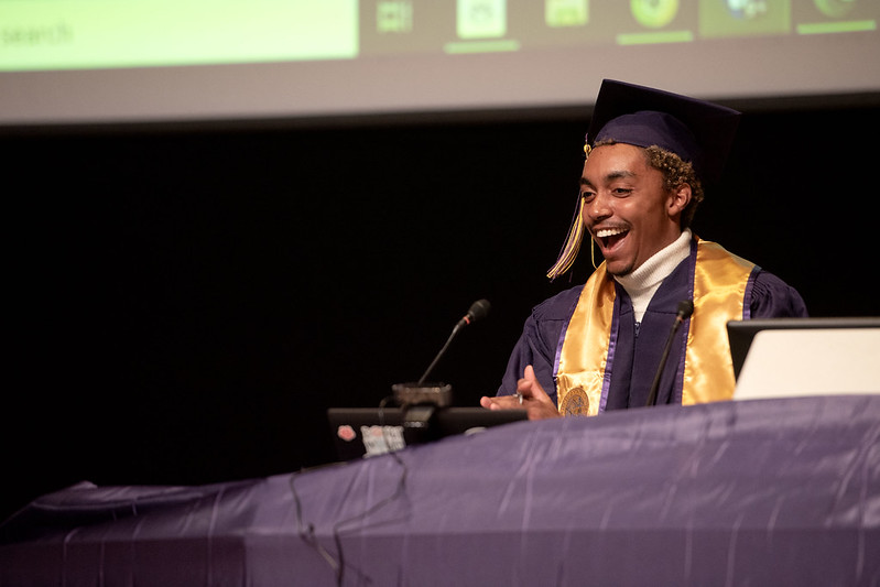 Troy Alexander smiles and looks to the left. He is giving a speech, and is wearing a purple graduation robe and cap, as well as a yellow graduation stole and tassel. He stands behind a podium.